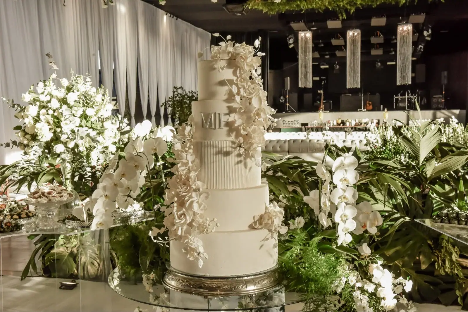 Bolo branco de vários andares cercado de orquídeas, rosas brancas e folhagens.