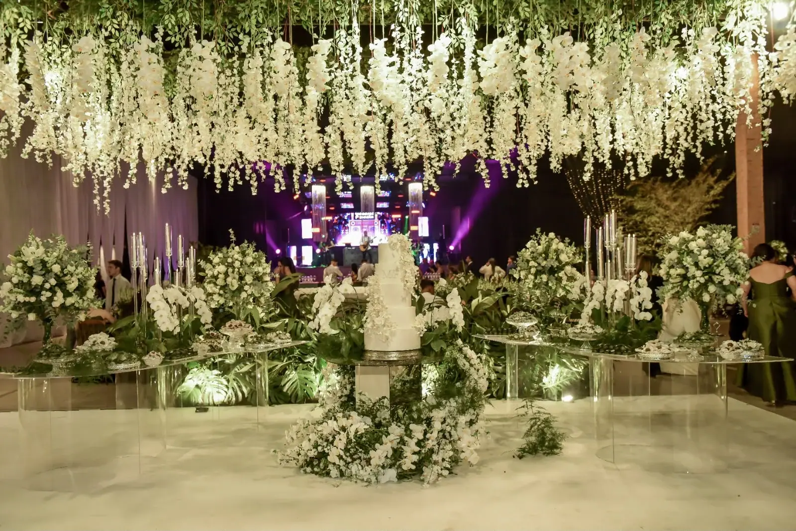 Mesa do bolo sob um teto de flores brancas pendentes, com arranjos, velas altas e o palco ao fundo.