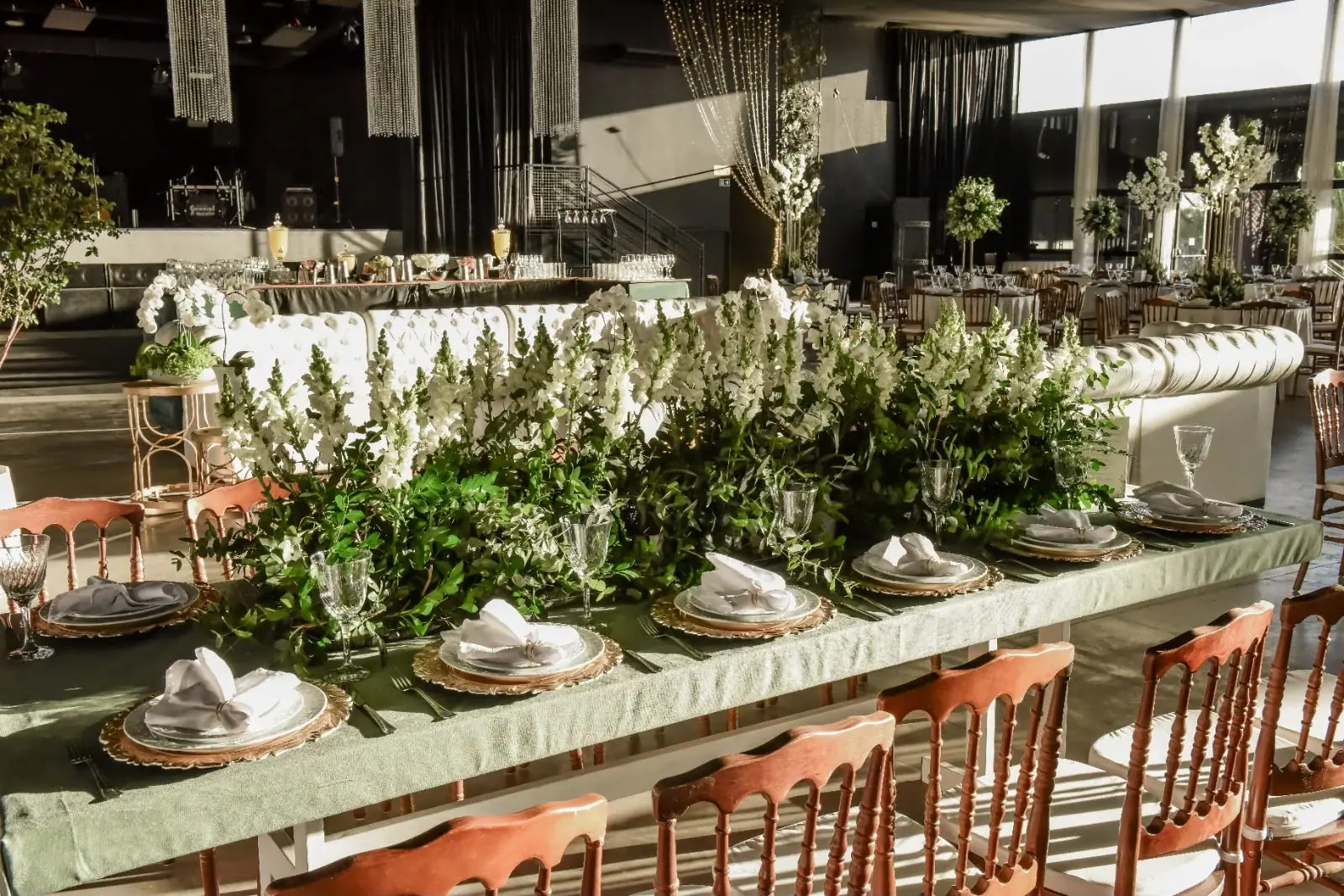 Mesa comprida com toalha verde, folhagens no centro e cadeiras de madeira.