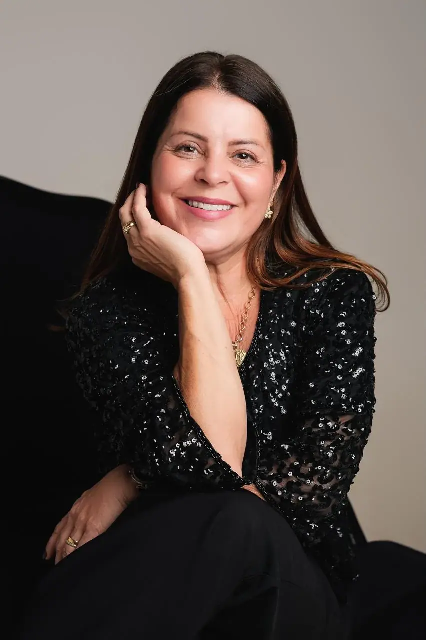 Valéria Moreira de blusa preta brilhante, sorrindo, com a mão no rosto.
