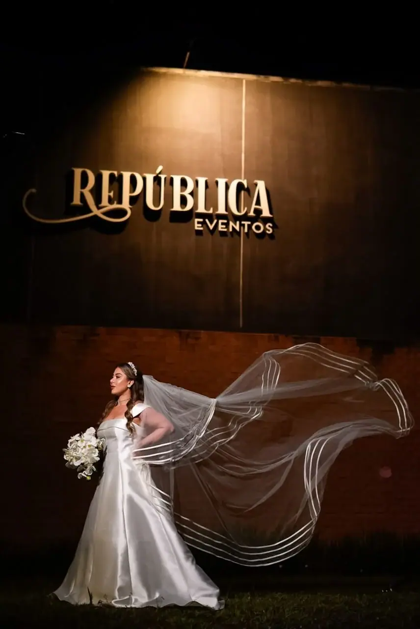 Noiva com o véu ao vento diante da fachada iluminada com o letreiro República Eventos.