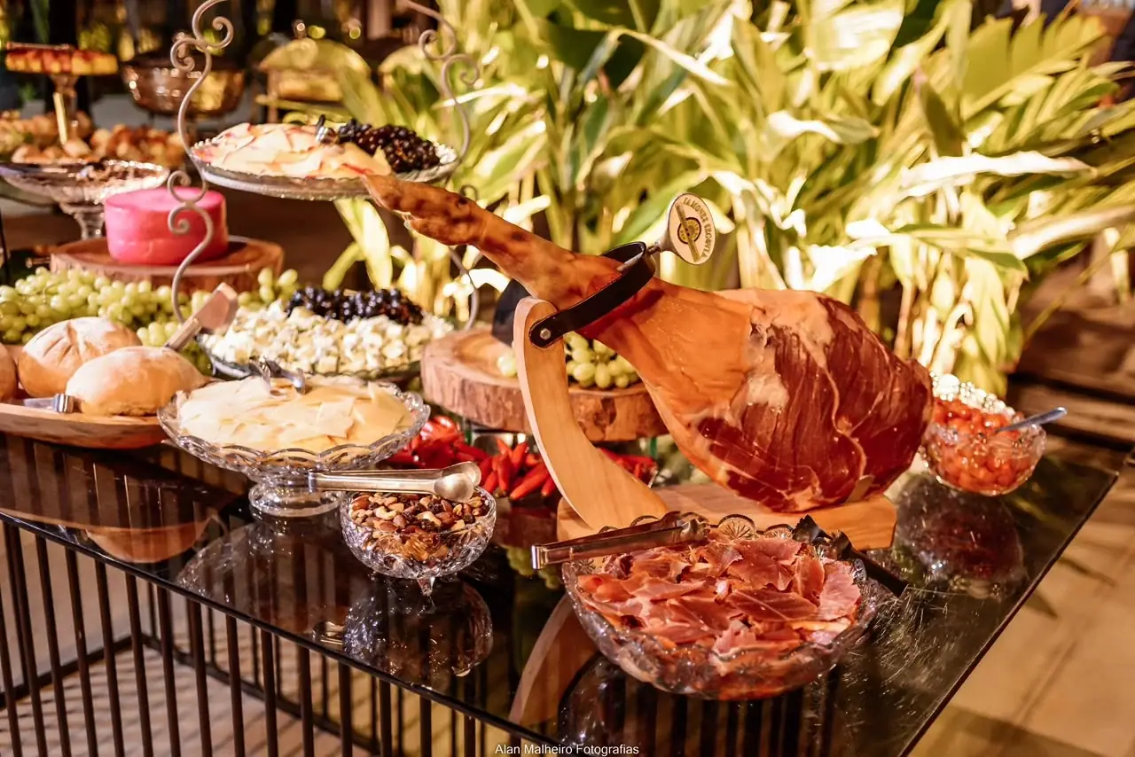 Mesa de frios com presunto cru, queijos, pães e frutas entre folhagens.