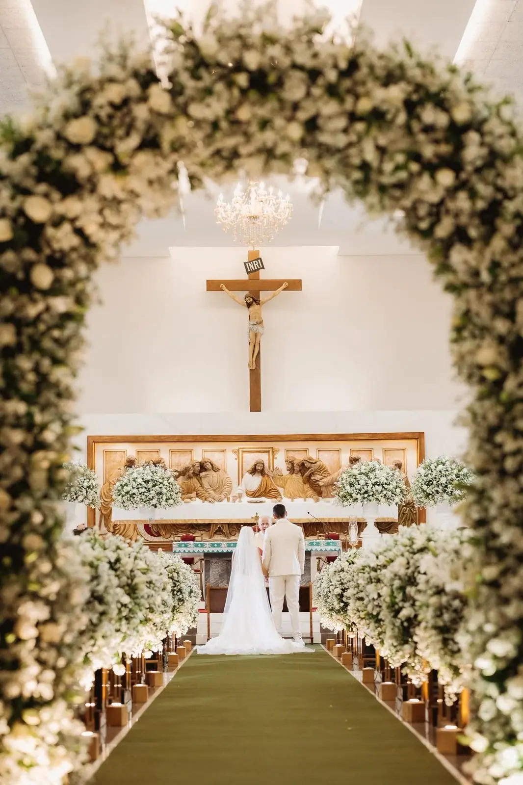 Corredor de igreja visto através de um arco de flores brancas, com os noivos no altar.